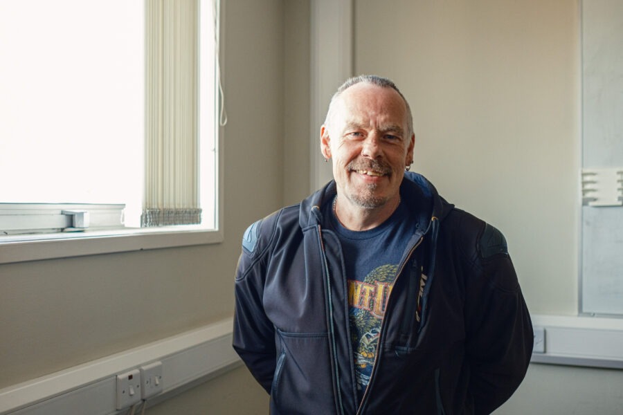 Alex, a man wearing a biker's jacket, standing smiling against a neutral background office environment.