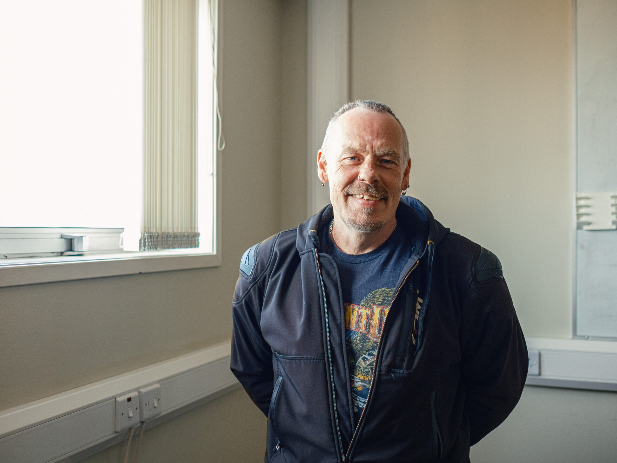 Alex, a man wearing a biker's jacket, standing smiling against a neutral background office environment.